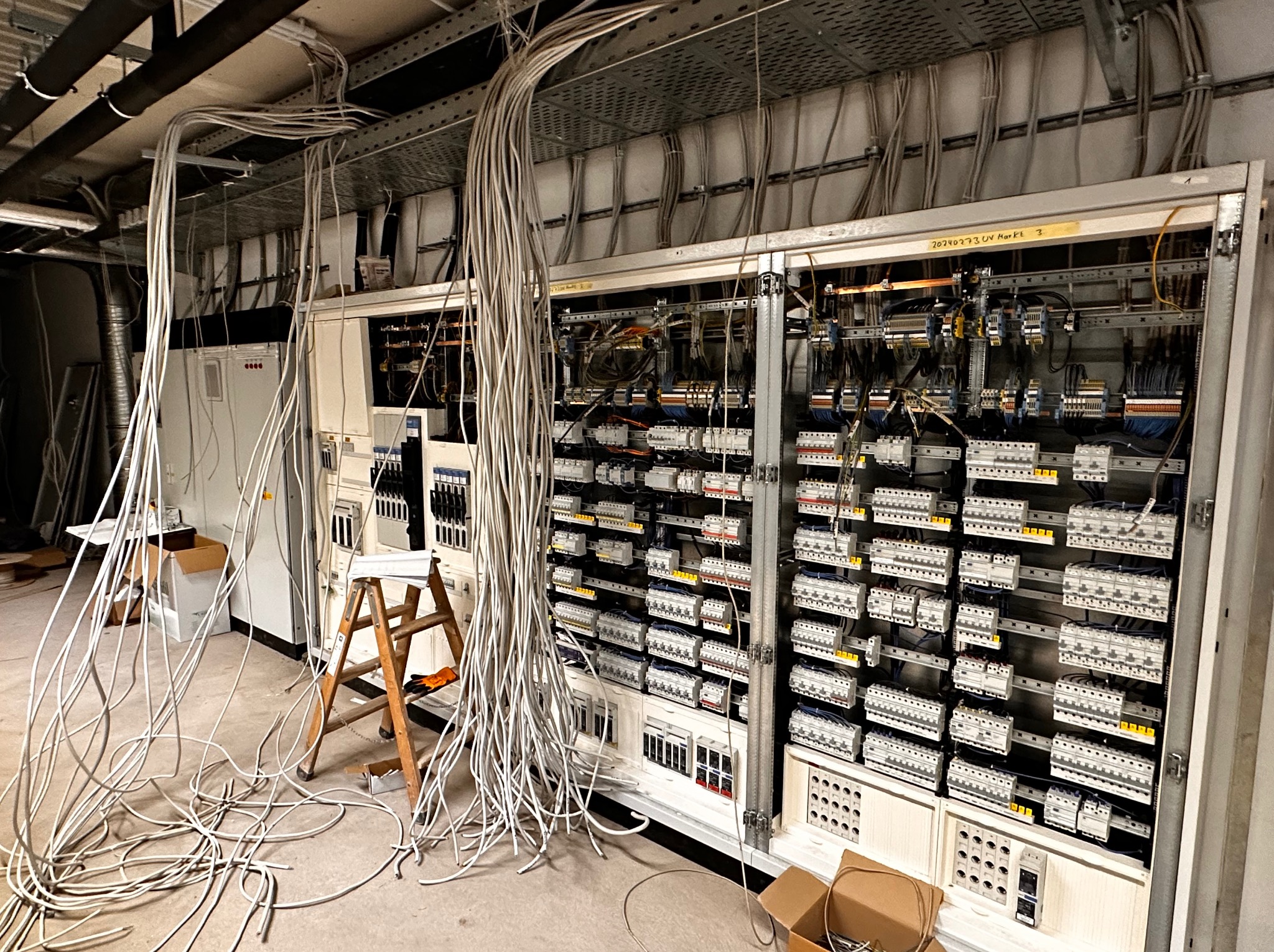 Rozbudowana szafa elektryczna z licznymi zabezpieczeniami i przewodami, widoczne luźno zwisające kable i drabina, w tle puszki i skrzynki elektryczne, surowy stan wykończenia.