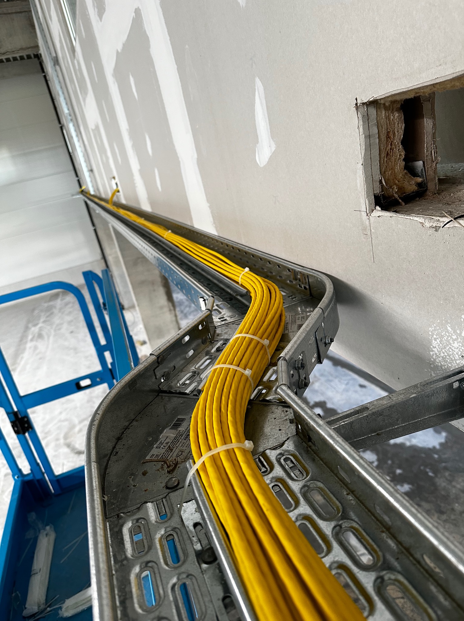 Żółte przewody elektryczne ułożone w metalowej korycie kablowej, zamocowanej do ściany w surowym stanie, widoczne elementy rusztowania.