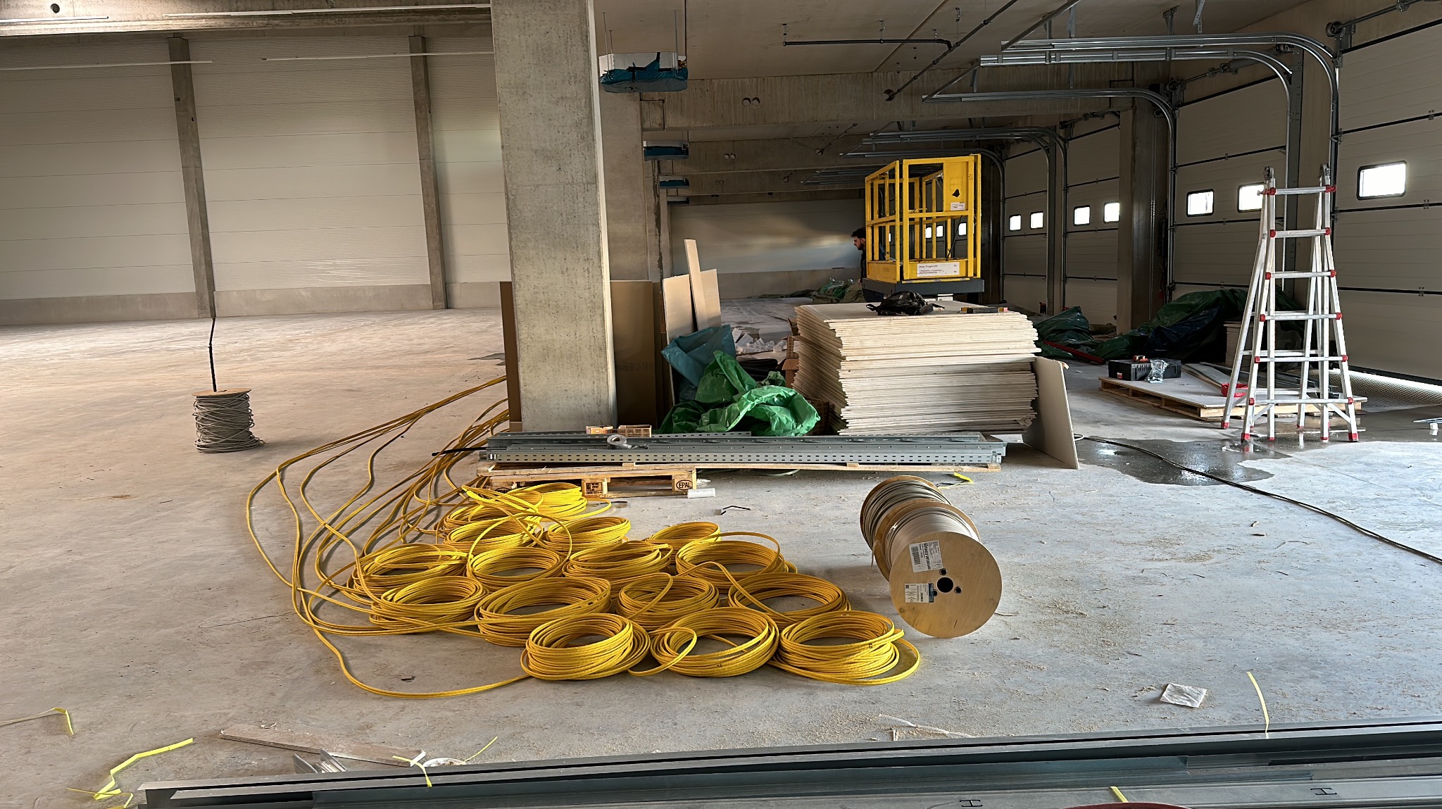 Wnętrze hali w trakcie prac wykończeniowych, widoczne zwitki żółtych kabli, szpule z przewodami, podnośnik koszowy i drabina.