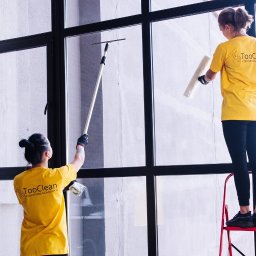 Komfort - Dwie kobiety w żółtych koszulkach z logo firmy czyszczą dużą, przeszkloną ścianę z czarną ramą, używając ściągaczki z długim uchwytem i białej rolki, jedna stoi na czerwonej drabinie.