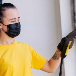 Komfort - Kobieta w żółtej koszulce i czarnej maseczce parowo czyści okno za pomocą parownicy z żółtymi elementami.