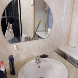 Komfort - Umywalka z kranem i drucianą myjką, w tle lustro owalne odbijające toaletę i mop oparty o ścianę, butelki detergentów na blacie.