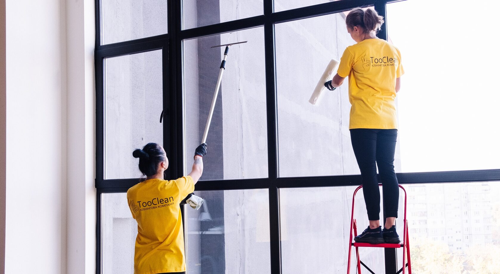 Dwie kobiety w żółtych koszulkach z logo firmy czyszczą dużą, przeszkloną ścianę z czarną ramą, używając ściągaczki z długim uchwytem i białej rolki, jedna stoi na czerwonej drabinie.