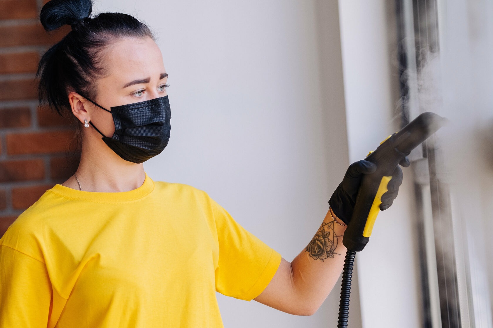 Kobieta w żółtej koszulce i czarnej maseczce parowo czyści okno za pomocą parownicy z żółtymi elementami.
