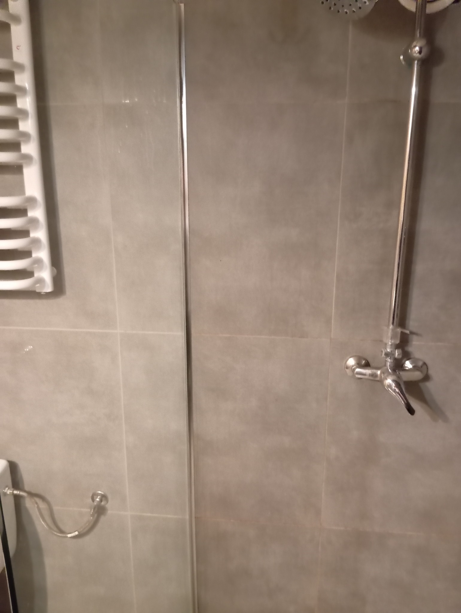 Szklana kabina prysznicowa z chromowaną armaturą i grzejnikiem drabinkowym na szarych płytkach.