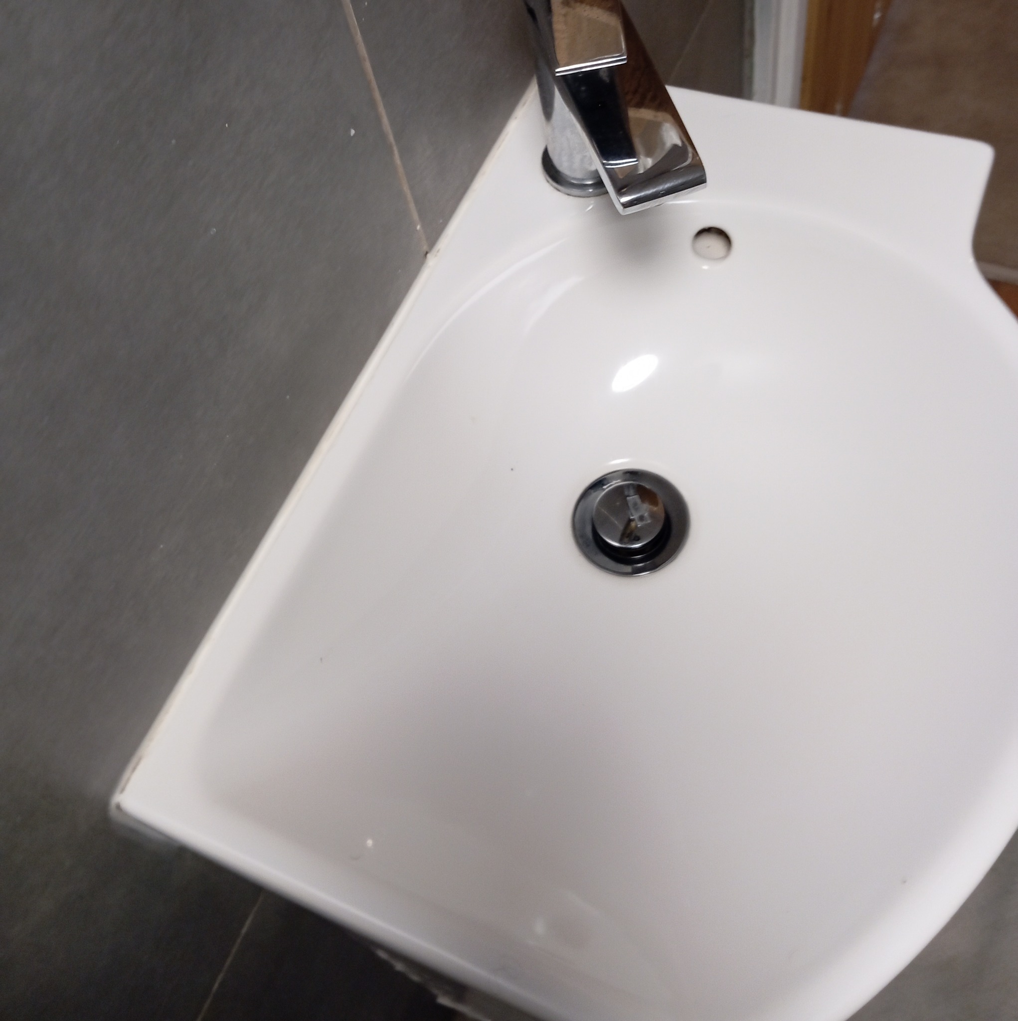 Biała, narożna umywalka z chromowaną baterią i odpływem, zamontowana na szarej ścianie z płytek.