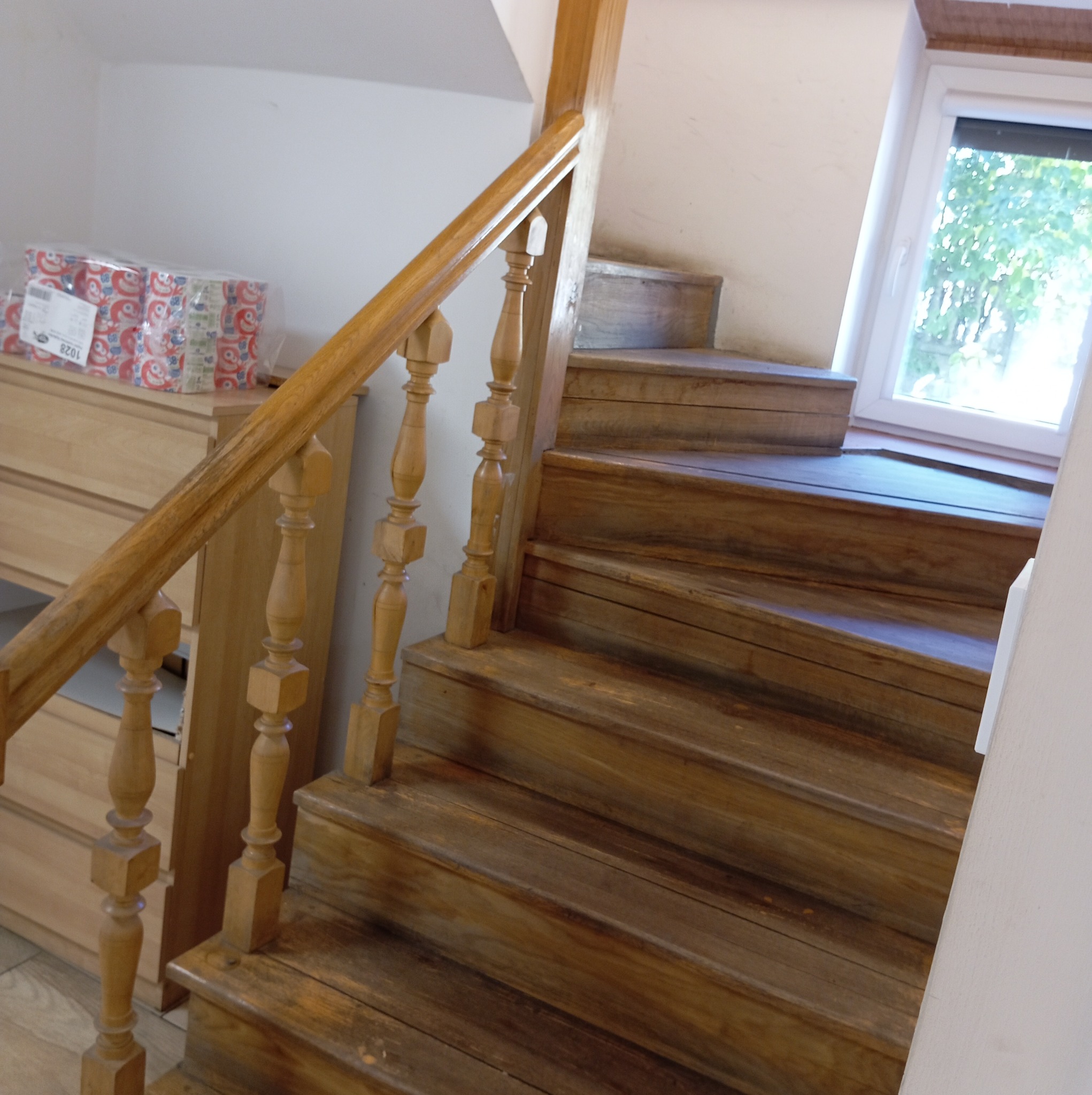 Drewniane schody z toczonymi balustradami w jasnym wnętrzu, widok z dołu, w tle komoda i okno z widokiem na zieleń.