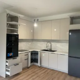 Jasna, nowoczesna kuchnia z beżowymi, błyszczącymi frontami, marmurkowym blatem i drewnianą podłogą. Widoczne otwarte szafki górne z mechanizmem podnoszenia, piekarnik w zabudowie, wysuwane...