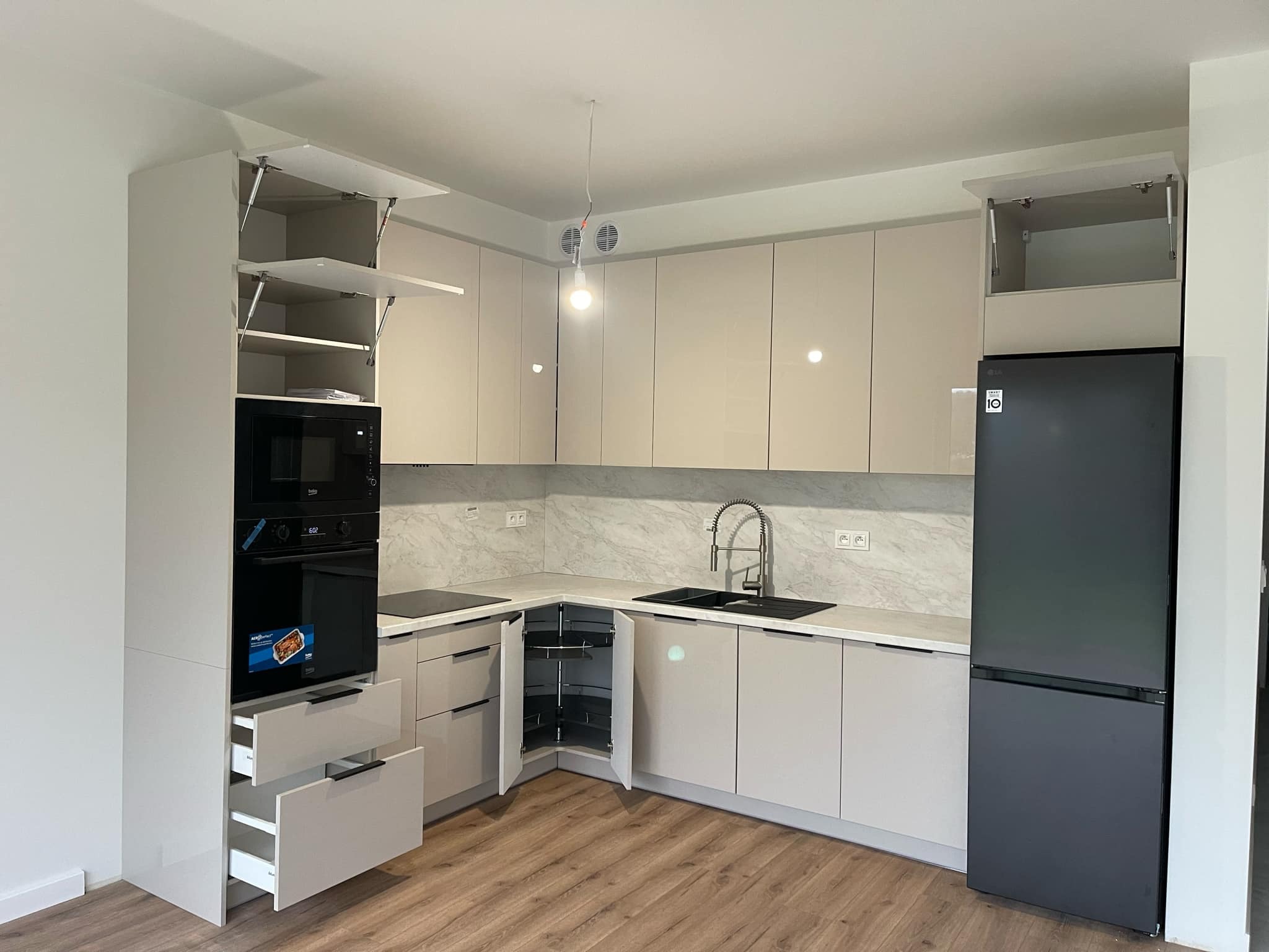 Jasna, nowoczesna kuchnia z beżowymi, błyszczącymi frontami, marmurkowym blatem i drewnianą podłogą. Widoczne otwarte szafki górne z mechanizmem podnoszenia, piekarnik w zabudowie, wysuwane...