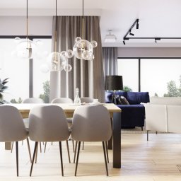 Zulo Design - Jasne wnętrze z drewnianą podłogą, stołem z szarymi krzesłami, szklanymi lampami wiszącymi, niebieską sofą i białym fotelem w tle, oświetlenie szynowe na suficie i projektor.
