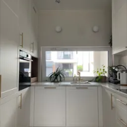 Biała kuchnia z drewnianą podłogą, złotymi uchwytami, roślinami w oknie i widokiem na budynek mieszkalny za oknem.