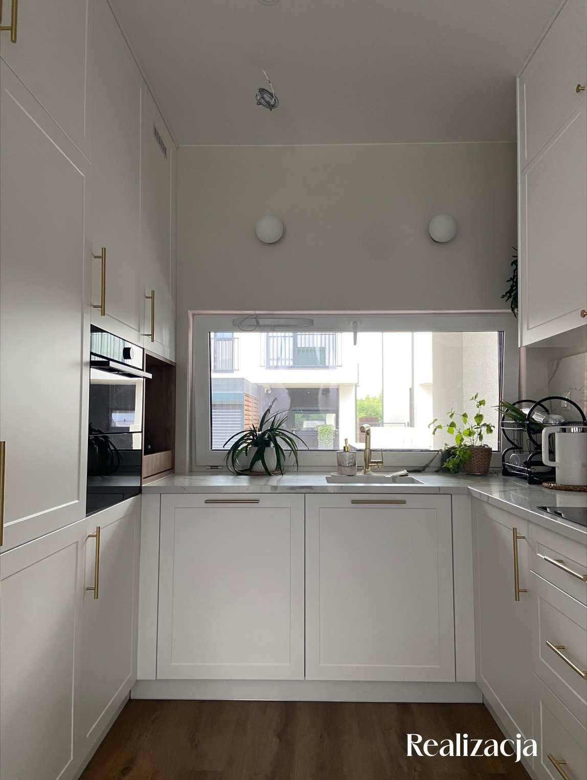 Biała kuchnia z drewnianą podłogą, złotymi uchwytami, roślinami w oknie i widokiem na budynek mieszkalny za oknem.
