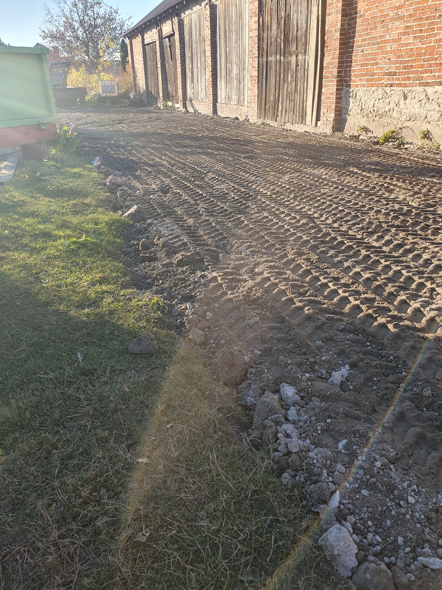 Świeżo wyrównany teren przy budynku gospodarczym z cegły, widoczne ślady opon maszyny budowlanej, trawa po lewej stronie, w tle drzewa bez liści i fragment zielonego kontenera.