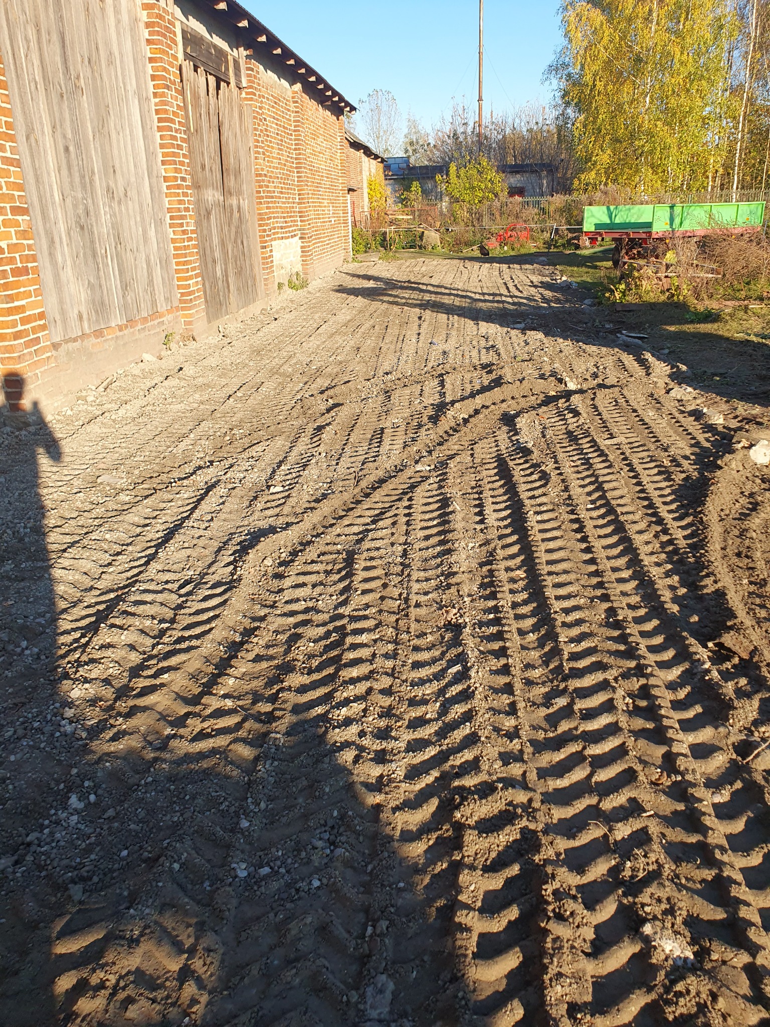 Świeżo utwardzona droga gruntowa z wyraźnymi śladami opon ciężkiego sprzętu, obok ceglanego budynku gospodarczego w słoneczny dzień.