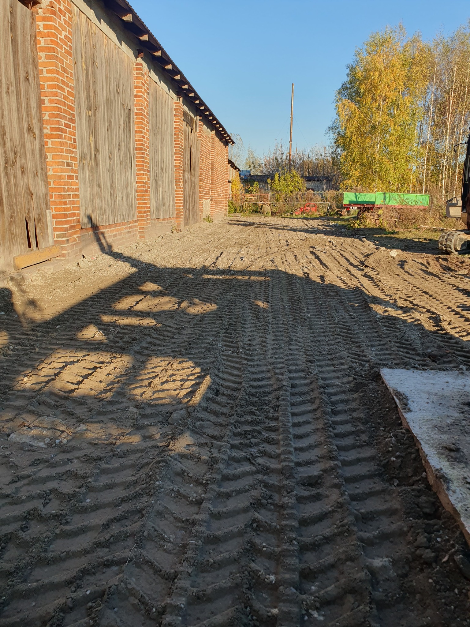 Przygotowany teren z widocznymi śladami opon maszyny budowlanej, ceglany budynek gospodarczy w tle, jesienne drzewa i fragment maszyny budowlanej po prawej stronie.