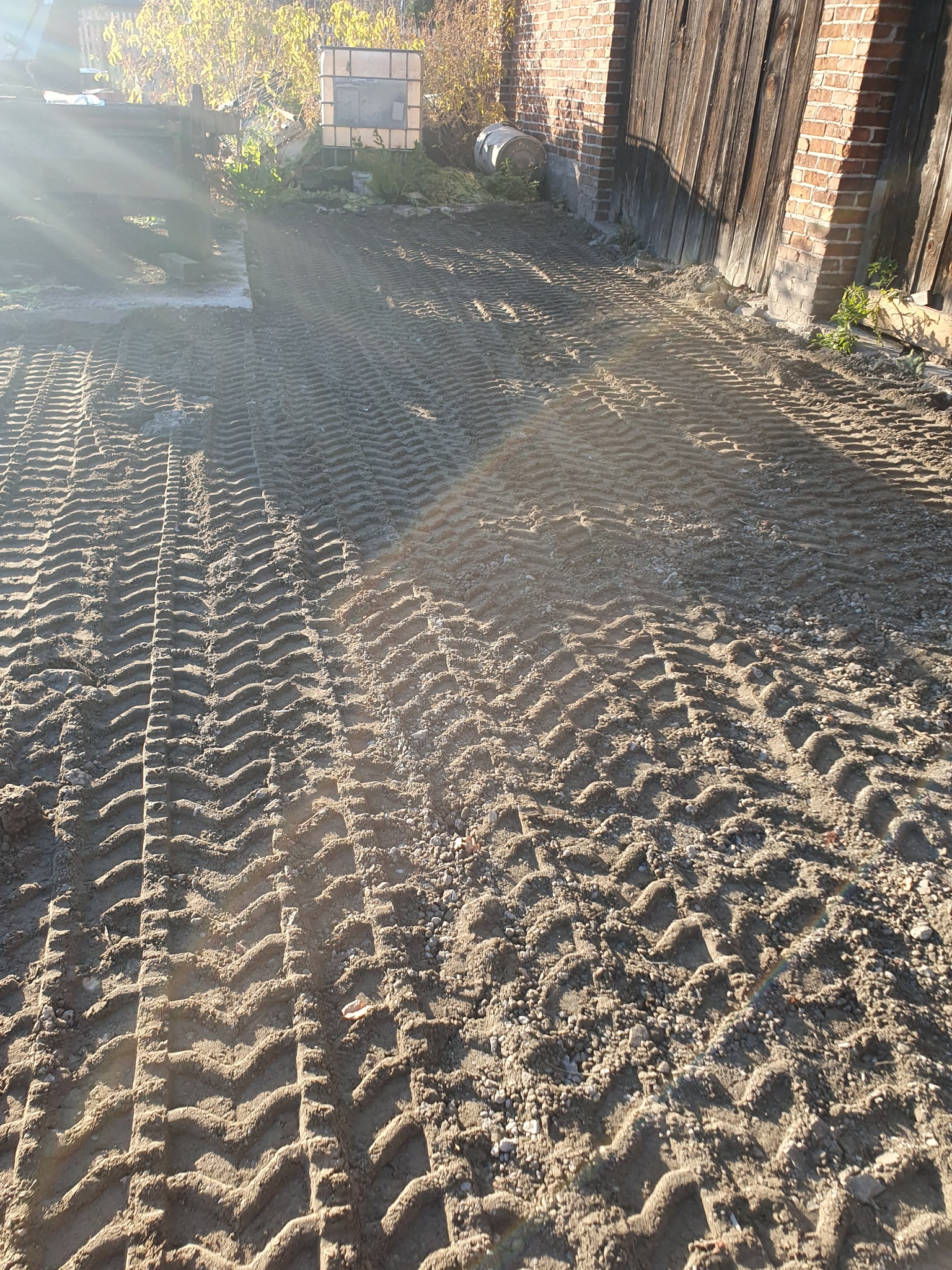 Świeżo wyrównany teren z widocznymi śladami opon maszyny budowlanej, w tle fragment budynku z cegły i drewnianymi drzwiami, oświetlone porannym słońcem.