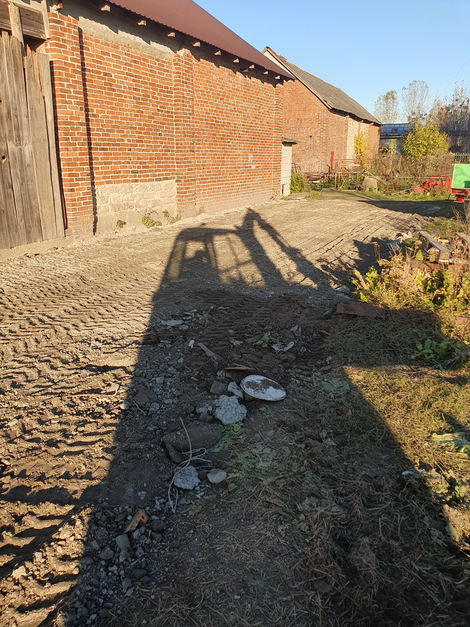 Cień koparki na nierównym, utwardzonym terenie z fragmentami gruzu i roślinności, w tle ceglany budynek gospodarczy w słoneczny dzień.