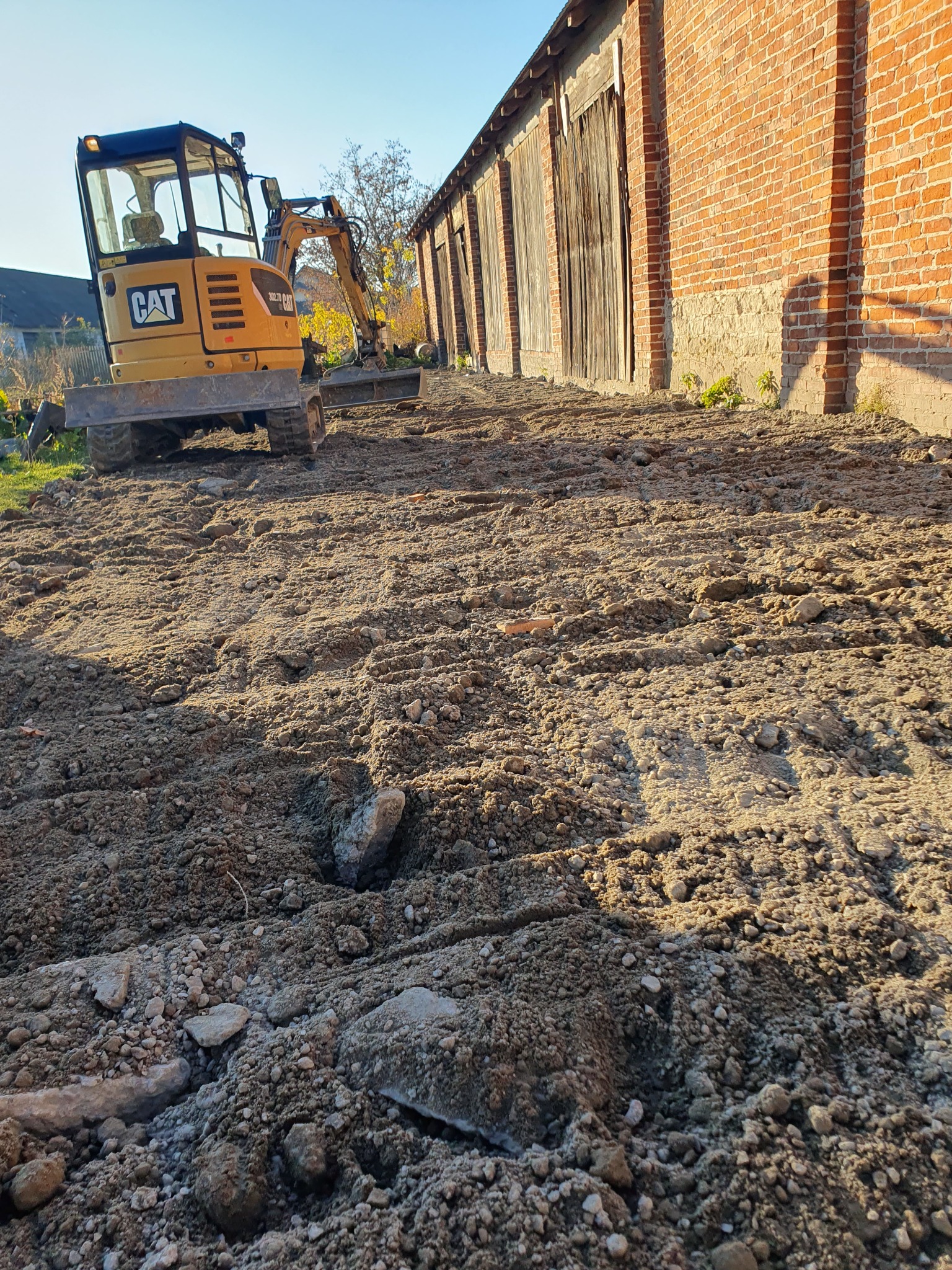 Żółta minikoparka CAT 302.7D na nierównym, częściowo wyrównanym terenie z widocznymi kamieniami, w tle ceglany budynek gospodarczy z drewnianymi wrotami, błękitne niebo.
