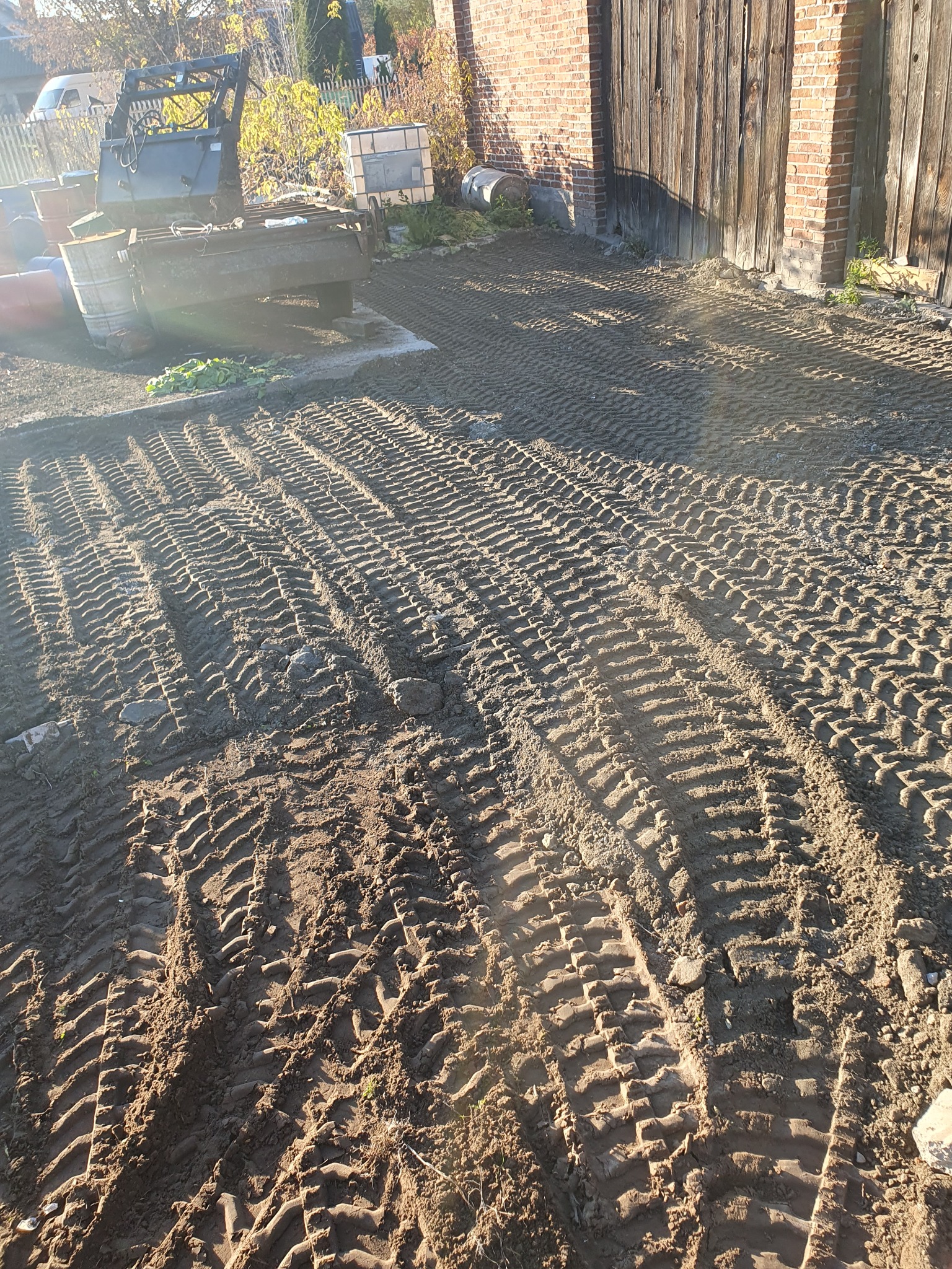 Świeżo wyrównany teren z wyraźnymi śladami opon ciężkiego sprzętu budowlanego, widoczne budynki w tle.