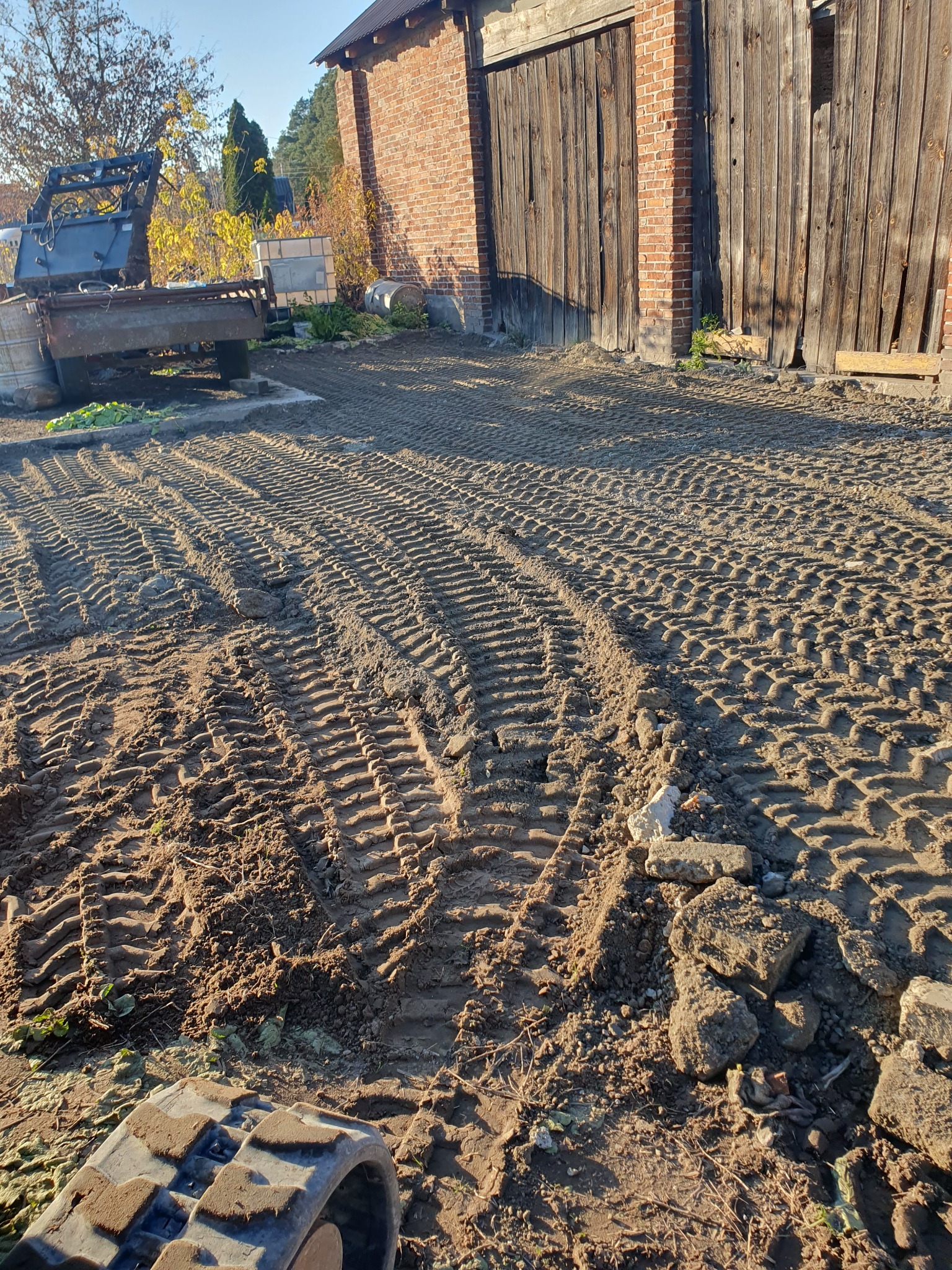Przygotowany, wyrównany teren pod budowę z widocznymi śladami gąsienic maszyny budowlanej, w tle budynek gospodarczy z drewnianymi wrotami i ceglaną ścianą.