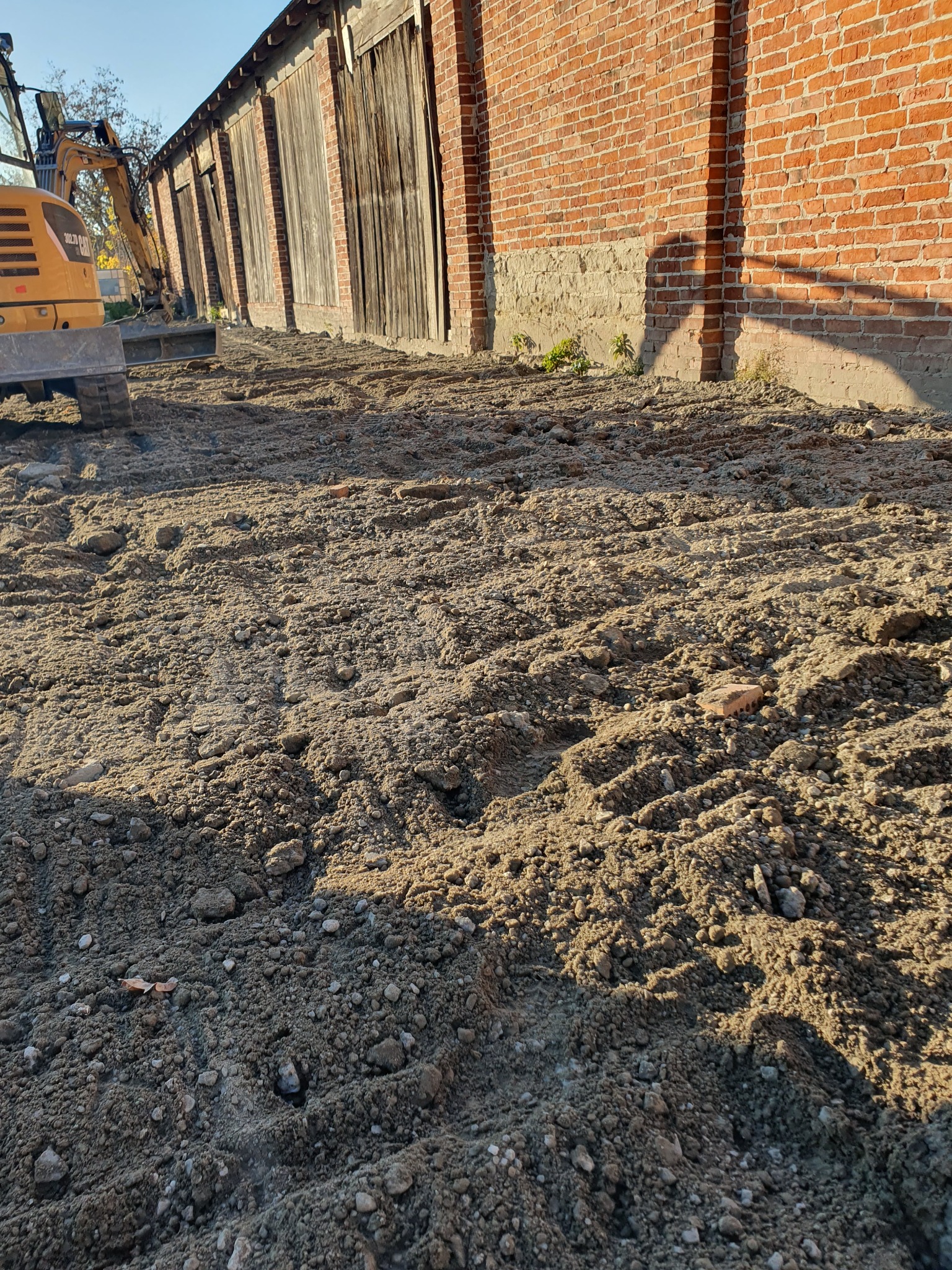 Żółta koparka Case CX30B na nierównym, częściowo utwardzonym gruncie z widocznymi śladami opon, obok ceglanego budynku z drewnianymi bramami w słoneczny dzień.