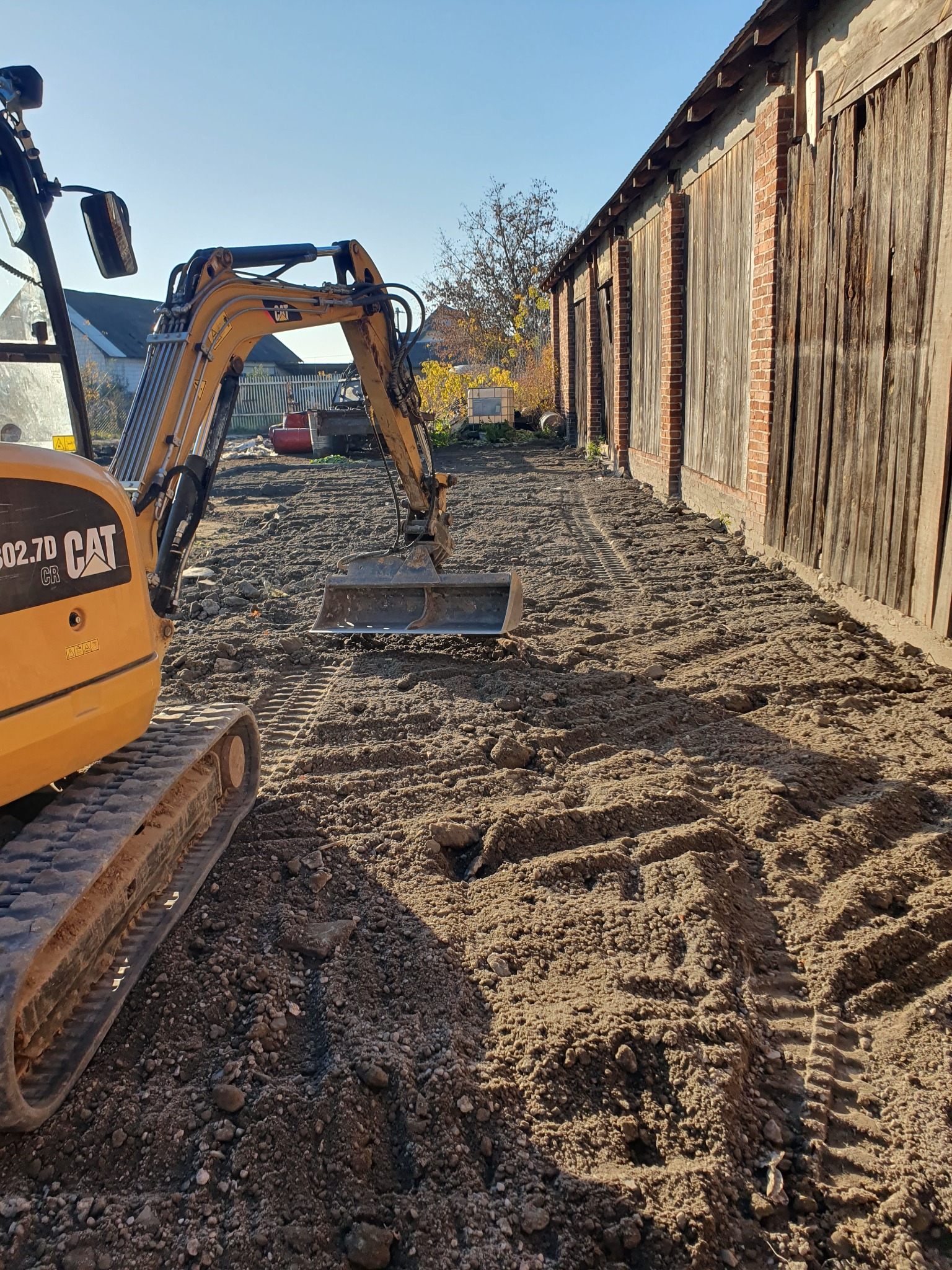 Żółta minikoparka CAT 302.7D CR na nierównym, brązowym terenie obok starego, drewniano-ceglanego budynku w słoneczny dzień.