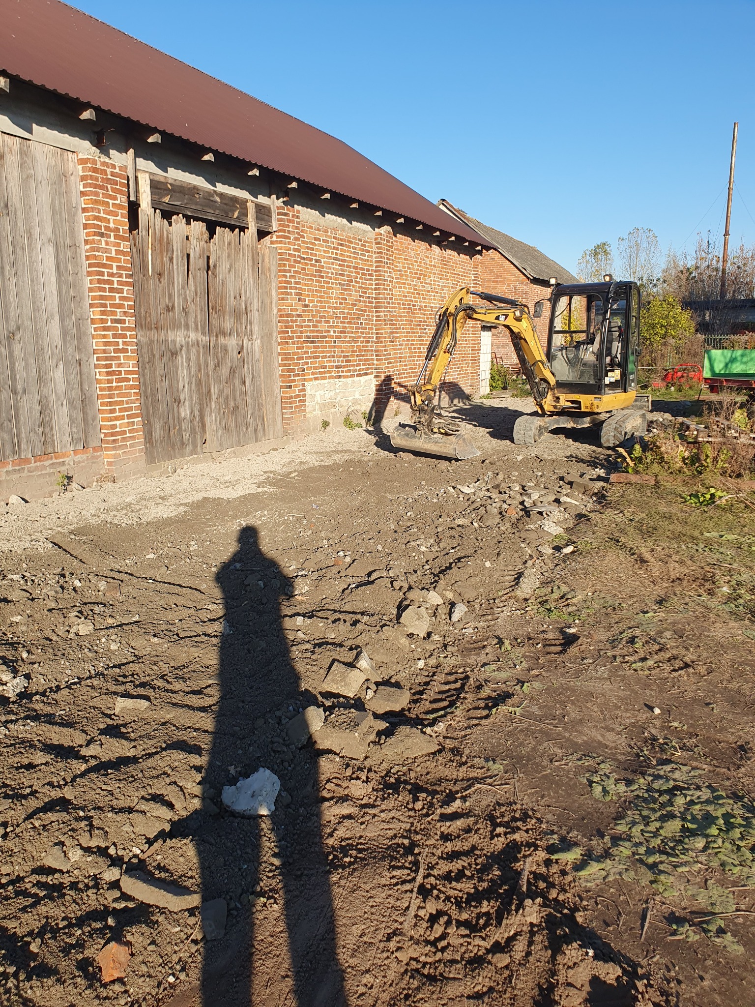 Żółta mini koparka na placu budowy przy ceglanym budynku gospodarczym z drewnianymi wrotami, widoczny cień osoby fotografującej.