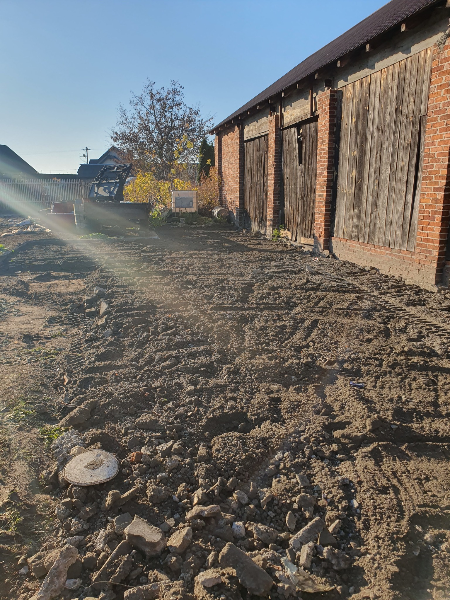 Przygotowany teren pod budowę z widoczną koparką, świeżo wyrównana ziemia z śladami opon, stare cegły w ziemi, budynek gospodarczy z cegły i drewnianymi wrotami w tle, jesienne drzewo i błękitne...
