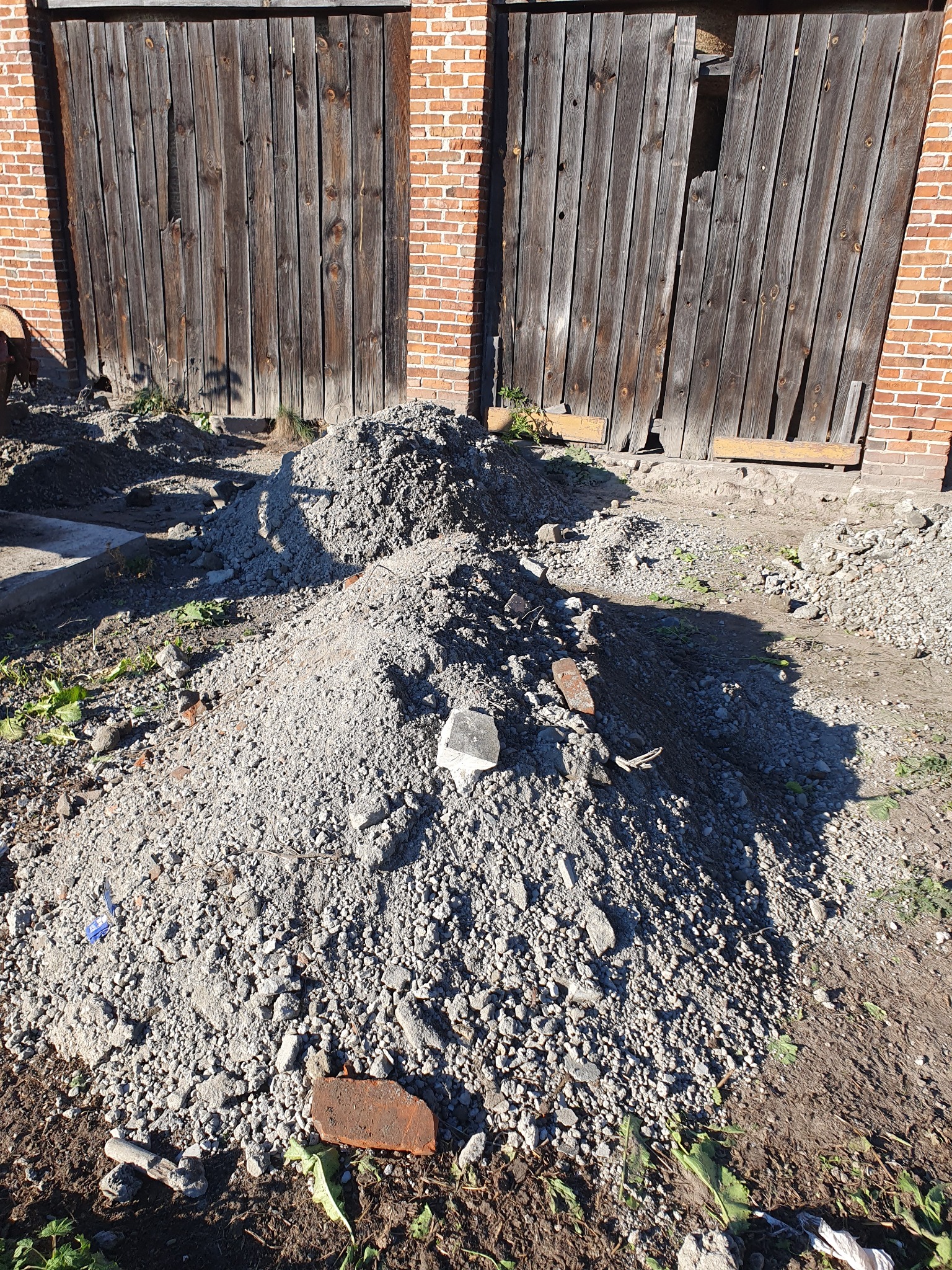 Dwie pryzmy gruzu i ziemi przed starą, ceglaną budowlą z drewnianymi, zniszczonymi wrotami, w tle ceglany budynek.