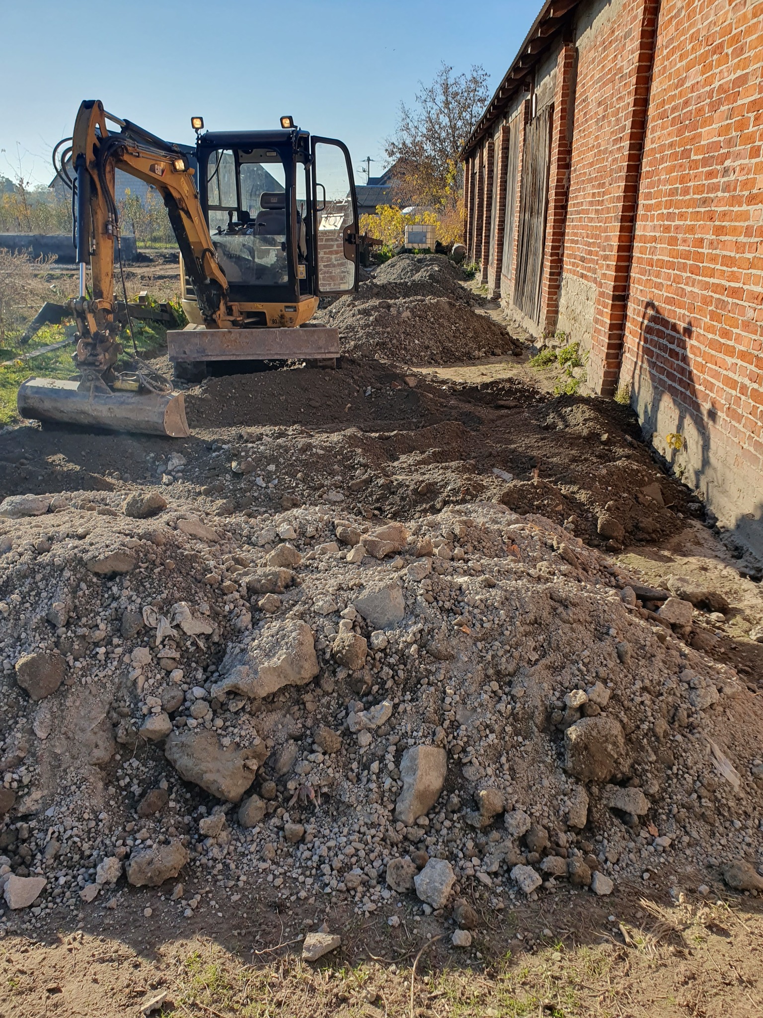 Żółta minikoparka CAT na placu budowy, z przodu pryzma ziemi z kamieniami, w tle budynek z czerwonej cegły i wykopany rów.