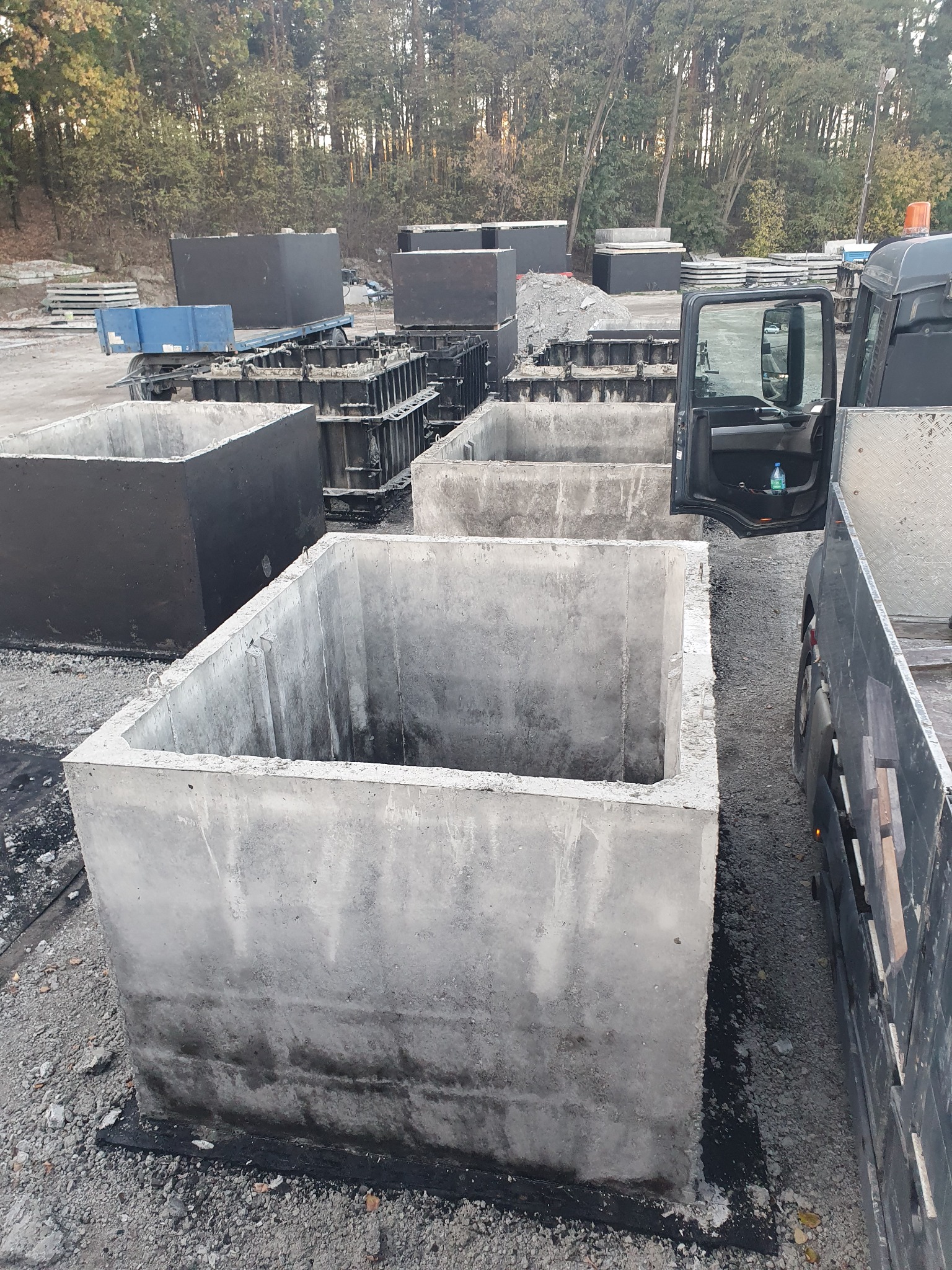 Betonowe, kwadratowe studzienki kanalizacyjne na placu budowy, widoczne formy do odlewania i samochód dostawczy z otwartymi drzwiami.