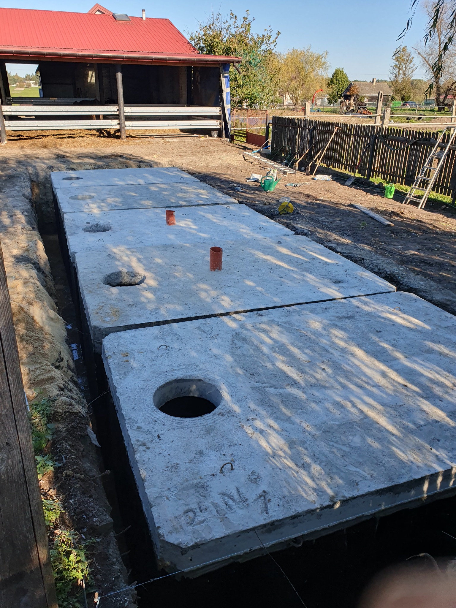 Prefabrykowane betonowe płyty szamba ułożone w wykopie, widoczne otwory rewizyjne i pionowe rury wentylacyjne, w tle budynek gospodarczy i ogrodzenie.
