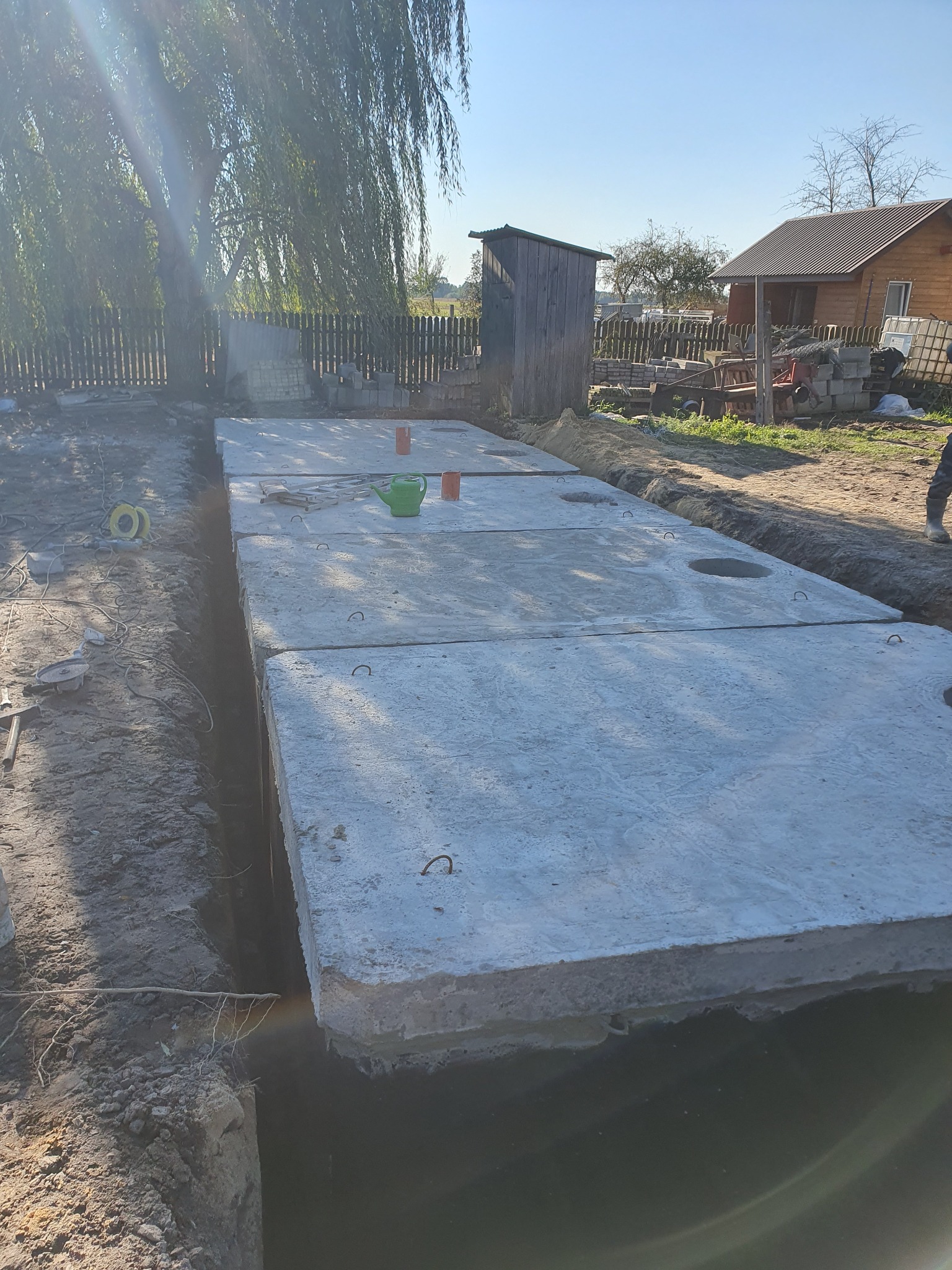 Betonowe płyty szamba ułożone w wykopie, widoczne haki do podnoszenia, na wierzchu zielony dzbanek i pomarańczowe kubki, w tle dom i drzewa.