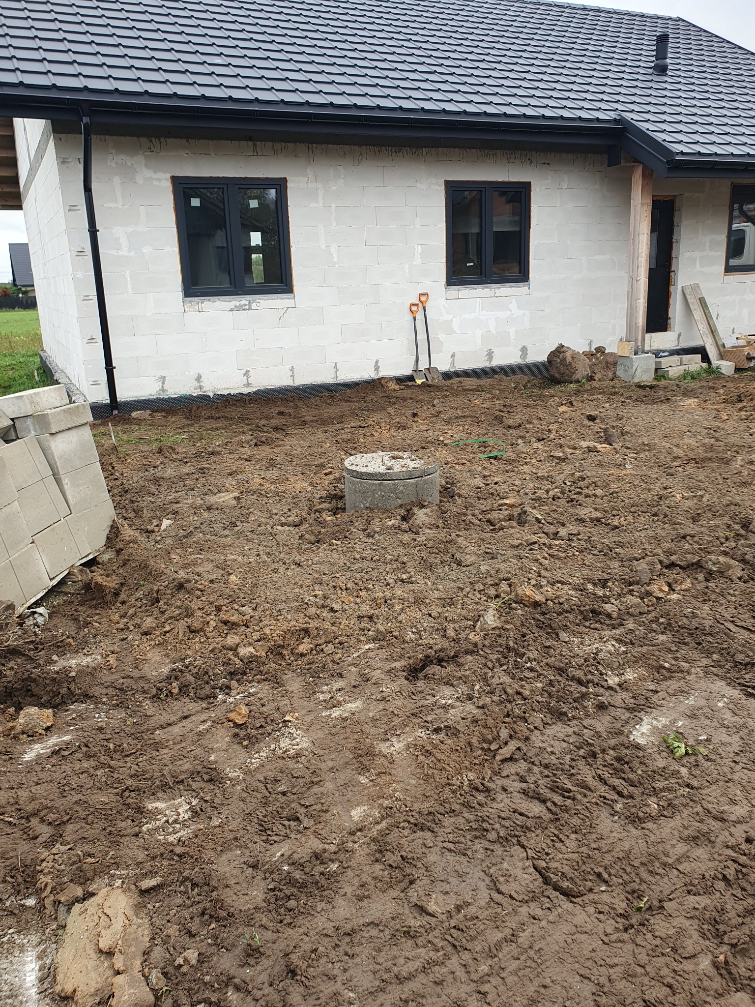 Teren po robotach ziemnych przy budowie domu jednorodzinnego, widoczne fundamenty i czarne okna, na ziemi leżą szpadle i betonowe kręgi