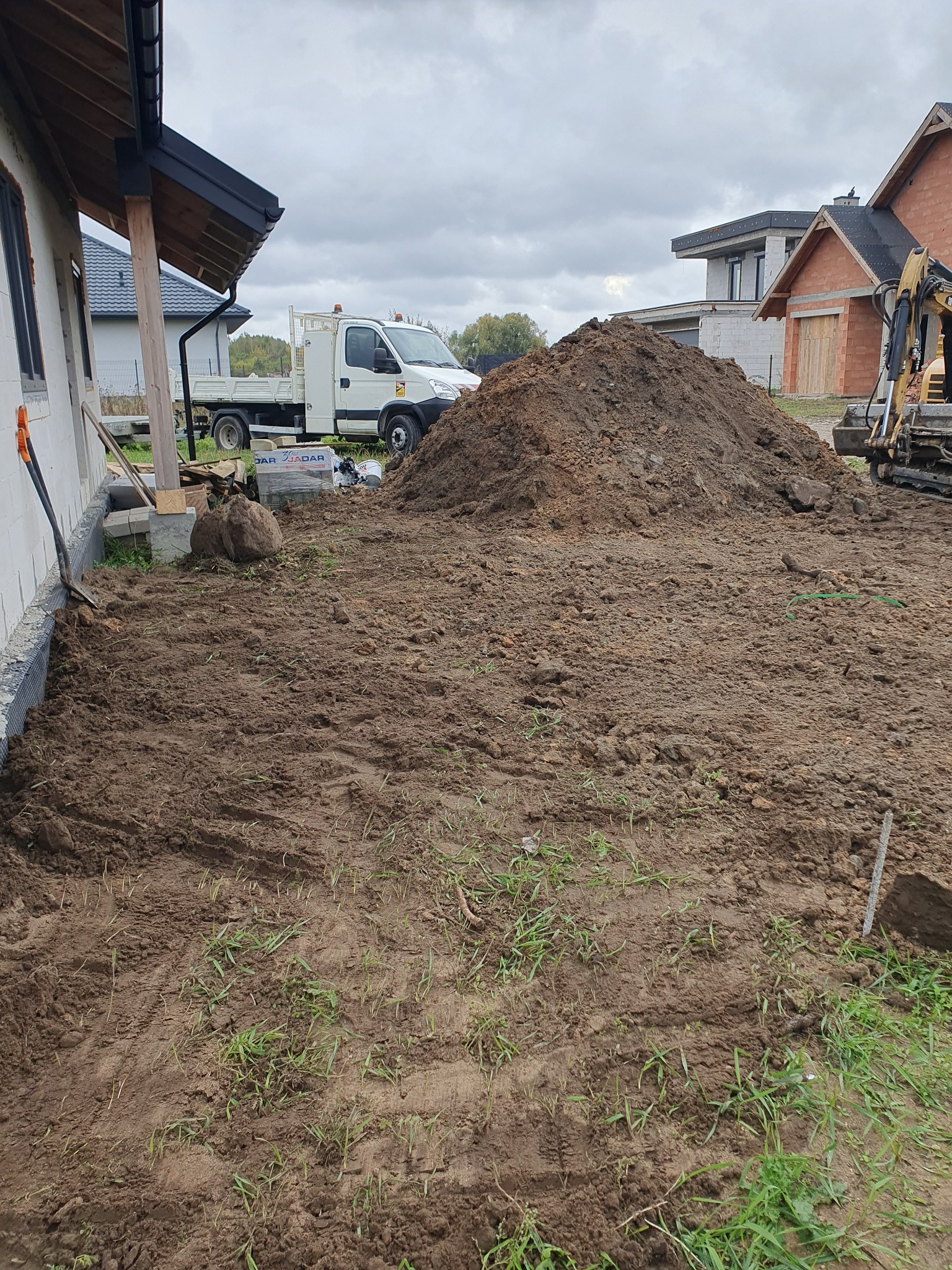 Przygotowany teren pod budowę z koparką, stertą ziemi, wykopem wzdłuż fundamentów i zaparkowanym samochodem dostawczym w tle.