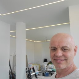 Zakład Usługowo Budowlany Grzegorz Gabliczak - Uśmiechnięty mężczyzna na tle białego wnętrza z oświetleniem LED w suficie, drabina i akcesoria malarskie w tle.