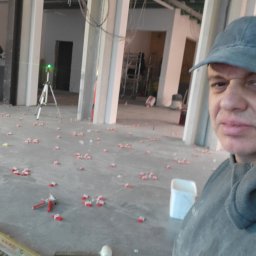 Zakład Usługowo Budowlany Grzegorz Gabliczak - Samodzielny montaż płytek podłogowych w surowym wnętrzu, widoczny laser krzyżowy, poziomica i przyssawka do płytek oraz rozstawione klipsy poziomujące, selfie wykonawcy.