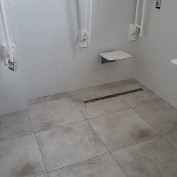 Zakład Usługowo Budowlany Grzegorz Gabliczak - Nowoczesna łazienka z beżowymi płytkami podłogowymi, białymi płytkami ściennymi, składanym siedziskiem i uchwytami dla osób niepełnosprawnych, widoczny odpływ liniowy.