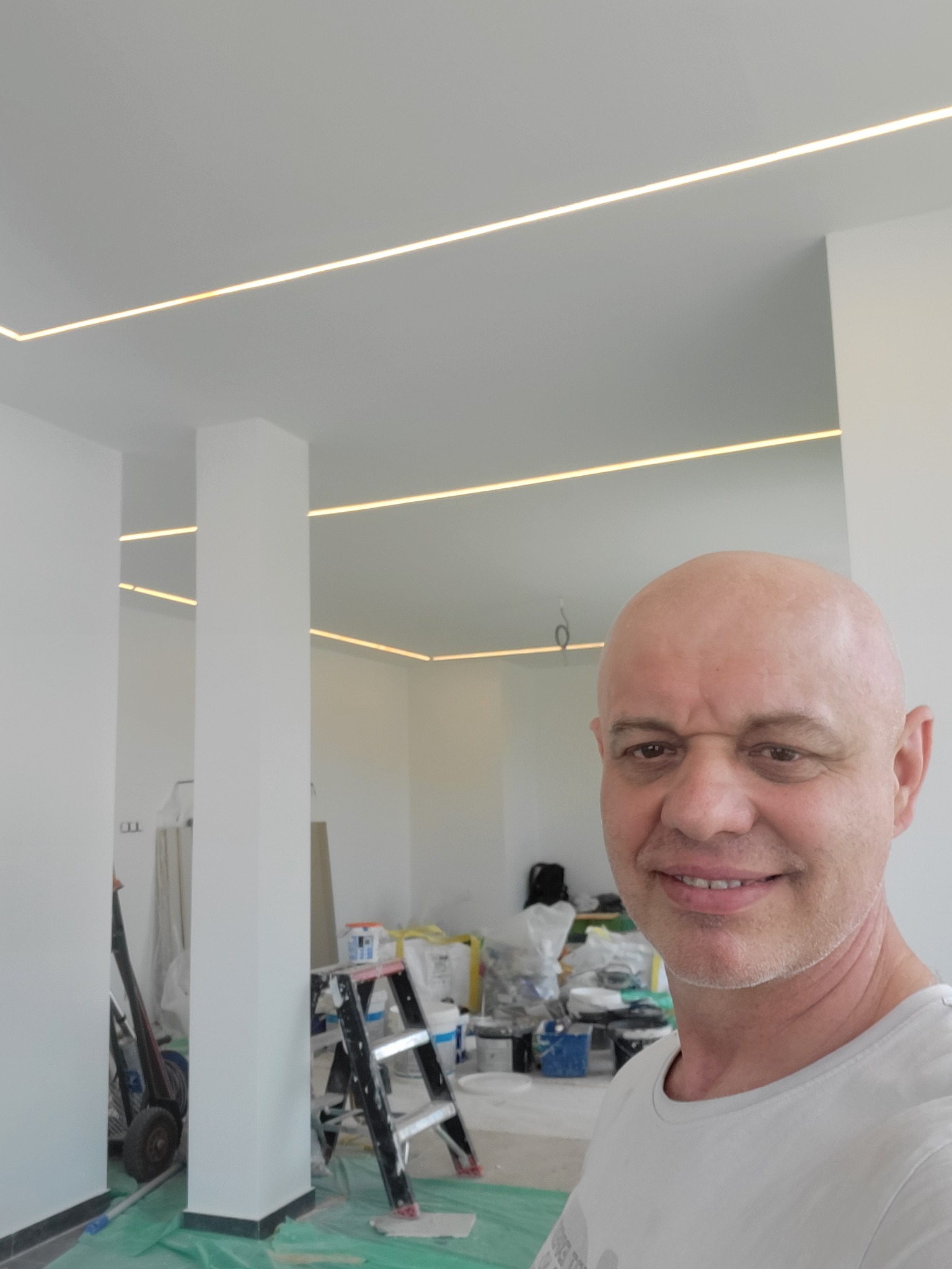 Uśmiechnięty mężczyzna na tle białego wnętrza z oświetleniem LED w suficie, drabina i akcesoria malarskie w tle.