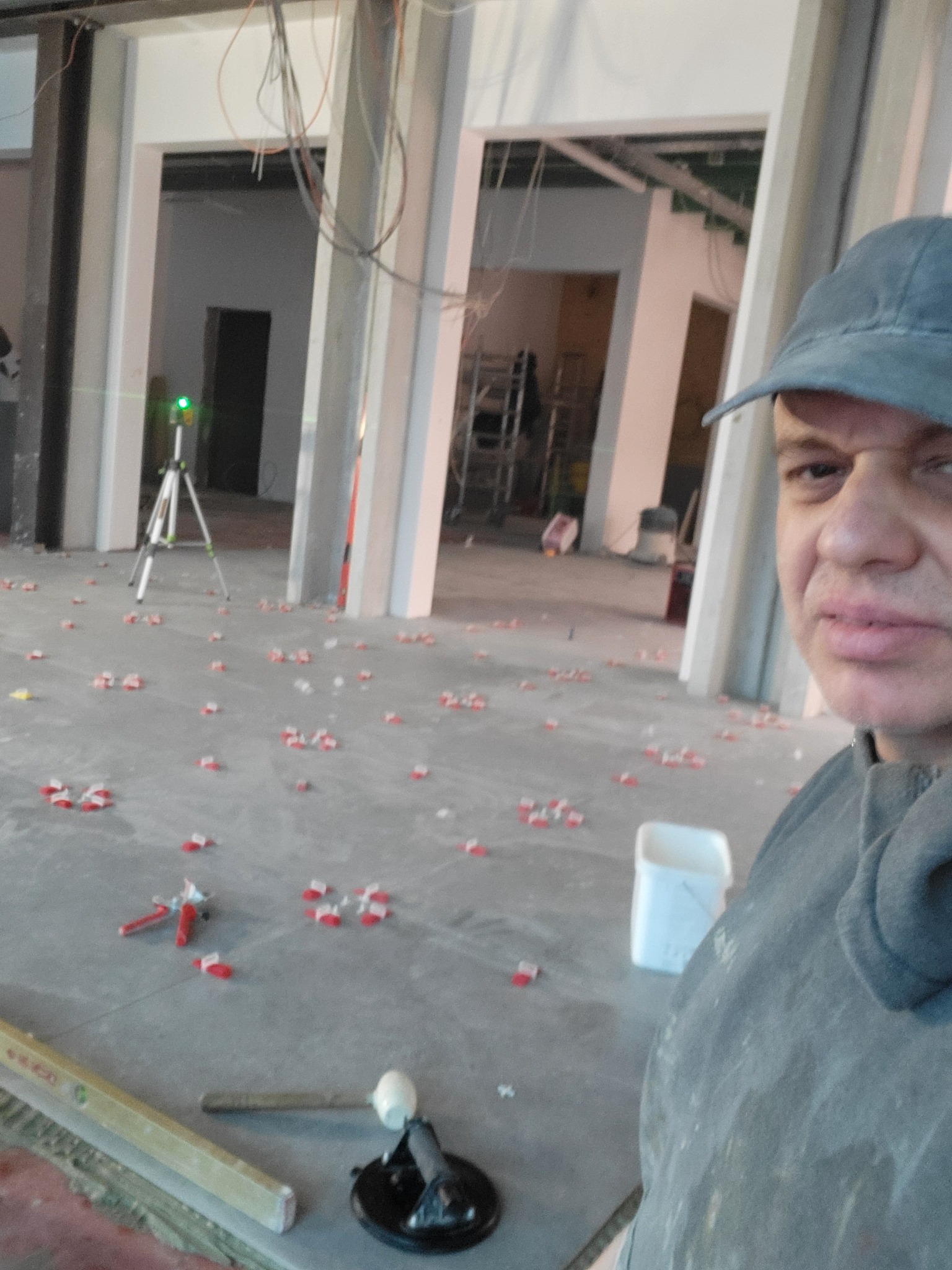 Samodzielny montaż płytek podłogowych w surowym wnętrzu, widoczny laser krzyżowy, poziomica i przyssawka do płytek oraz rozstawione klipsy poziomujące, selfie wykonawcy.