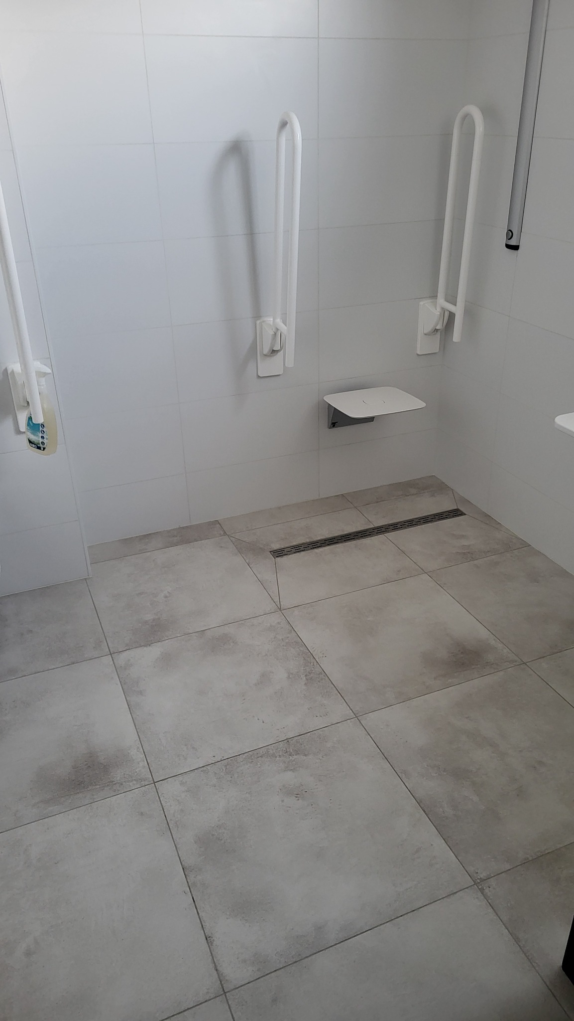 Nowoczesna łazienka z beżowymi płytkami podłogowymi, białymi płytkami ściennymi, składanym siedziskiem i uchwytami dla osób niepełnosprawnych, widoczny odpływ liniowy.