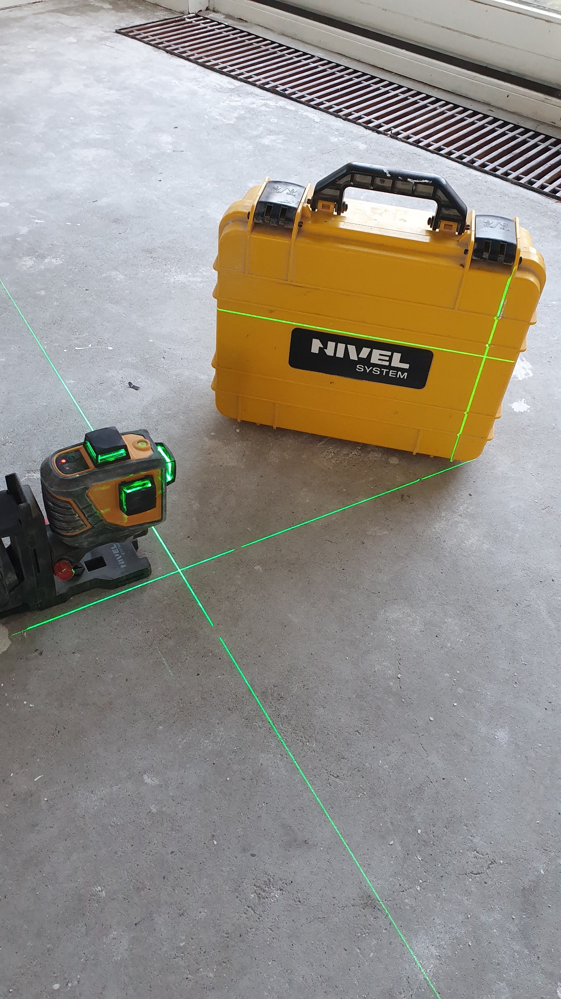 Poziomica laserowa emitująca zielone linie krzyżowe na szarej posadzce, obok żółta skrzynka transportowa marki Nivel System.