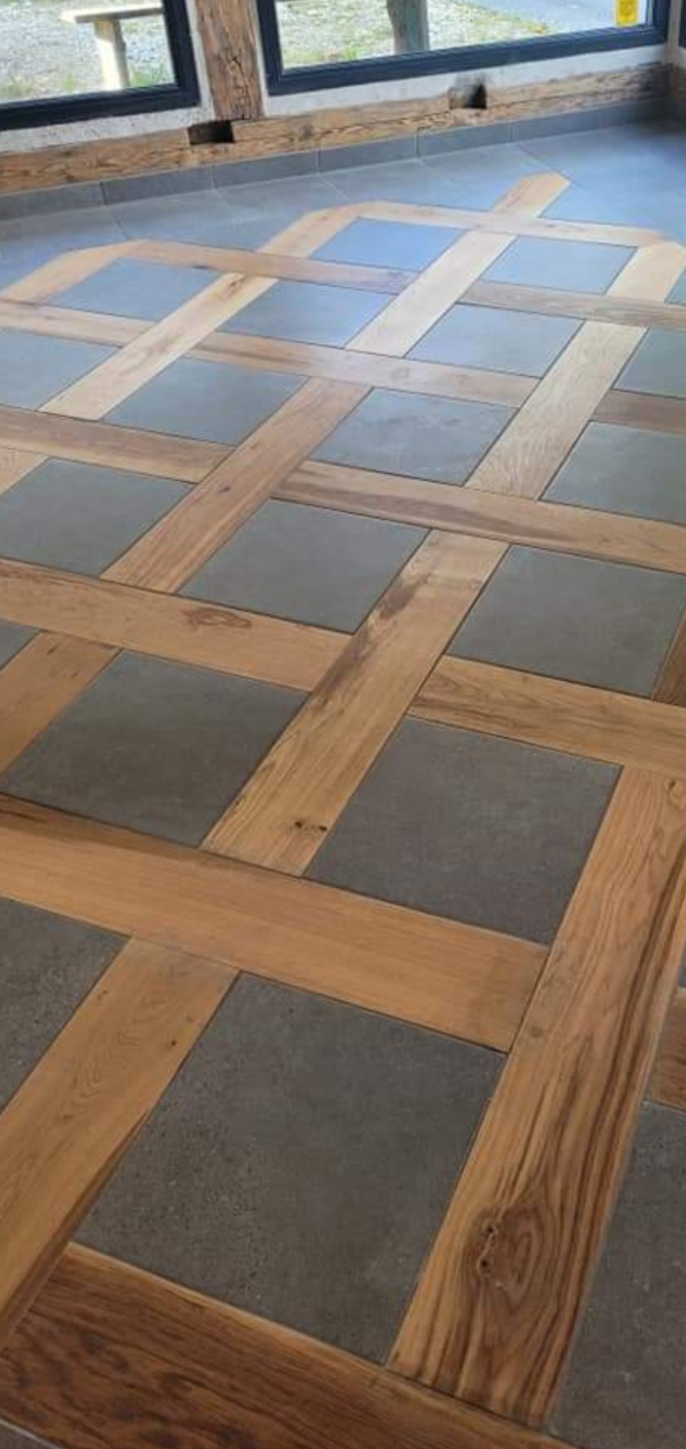 Dekoracyjna podłoga z płytek w odcieniach szarości, ułożonych w kwadraty, obramowanych jasnym drewnem w geometryczny wzór kraty.