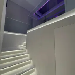 Nowoczesne, białe schody z podświetleniem LED na każdym stopniu, szklana balustrada, dodatkowe fioletowe oświetlenie w górnej części kadru.