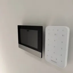 Panel dotykowy wideodomofonu Hikvision z klawiaturą numeryczną na białej ścianie, widok z boku.