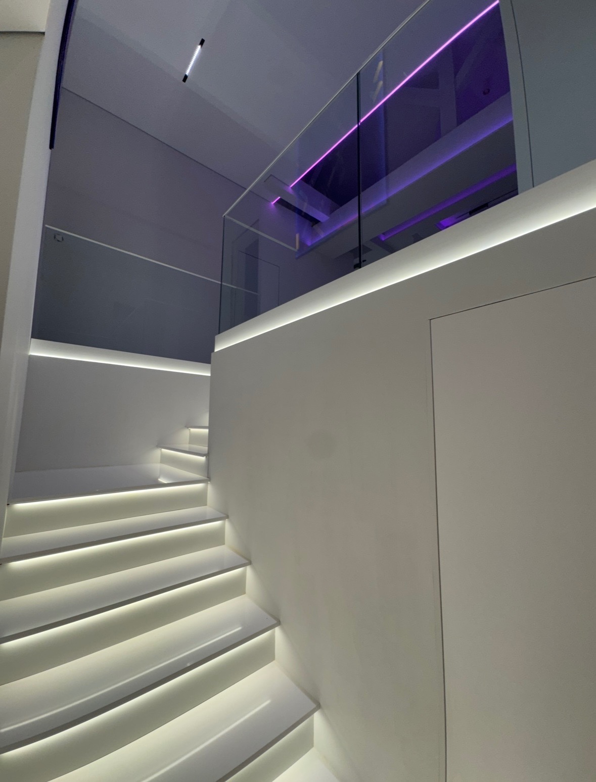 Nowoczesne, białe schody z podświetleniem LED na każdym stopniu, szklana balustrada, dodatkowe fioletowe oświetlenie w górnej części kadru.