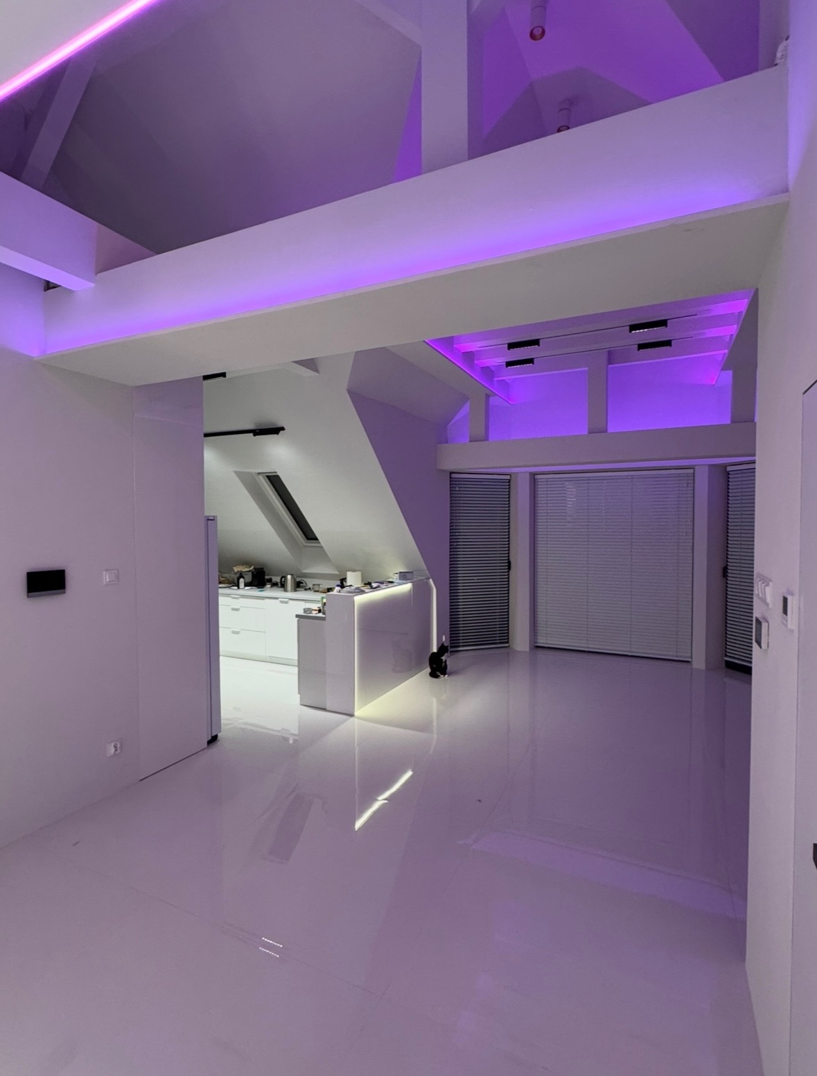 Nowoczesne, białe wnętrze z oświetleniem LED w kolorze fioletowym, widoczna kuchnia w tle, minimalistyczny design.