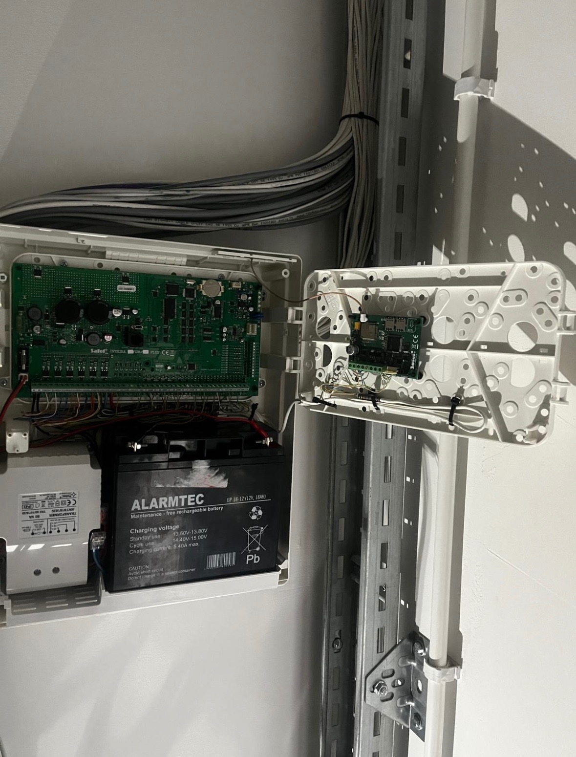 Otwarta obudowa centrali alarmowej z widoczną elektroniką, akumulatorem ALARMTEC i wiązką kabli u góry, zamontowana na szynie DIN.