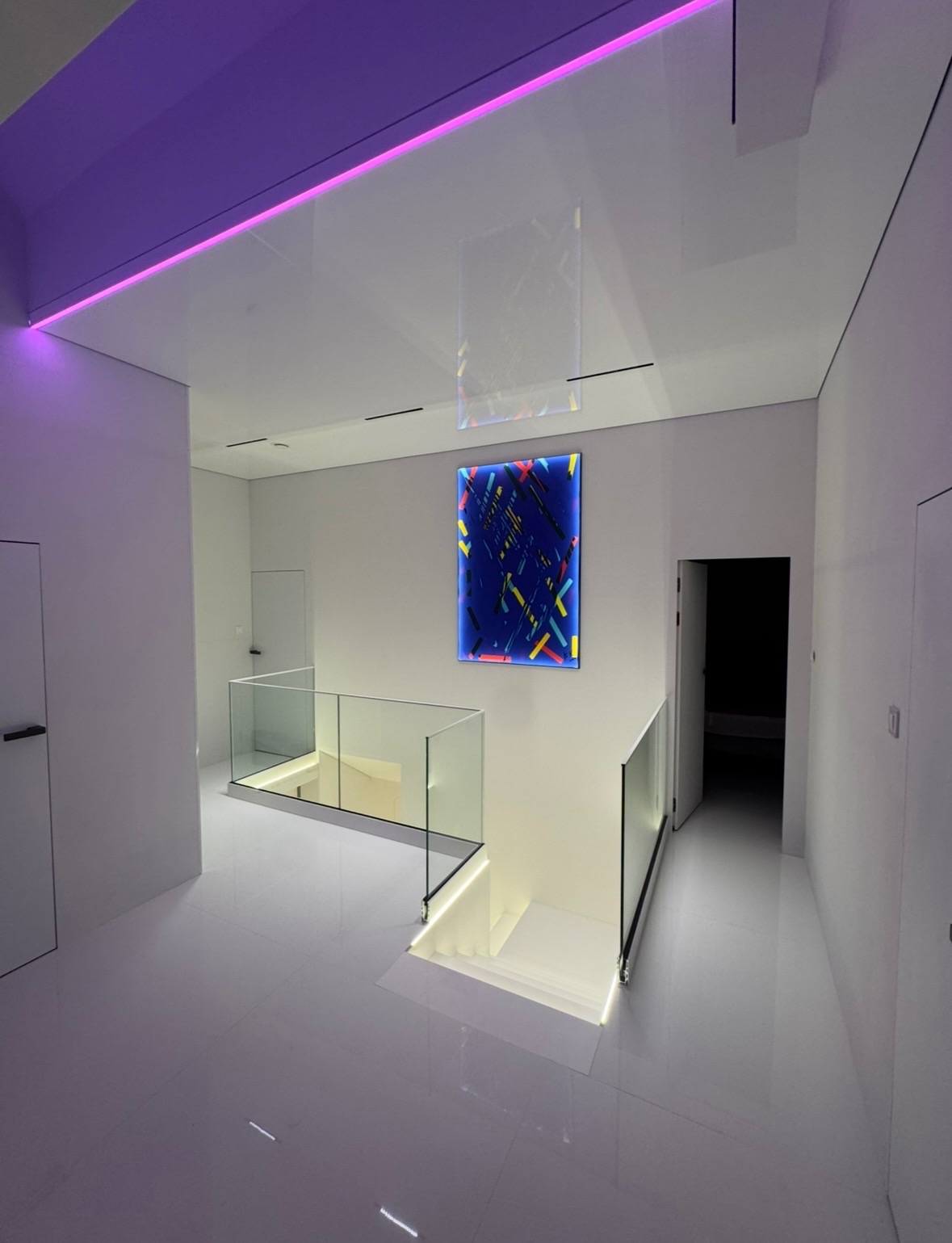 Nowoczesne wnętrze z białymi ścianami i podłogą, szklane balustrady schodów, podświetlenie LED w kolorze fioletowym na suficie i niebieski panel dekoracyjny z kolorowymi paskami na ścianie.