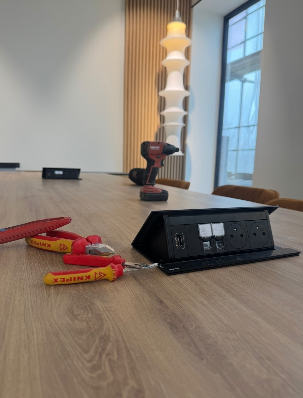Wpuszczana listwa zasilająca z gniazdami elektrycznymi, HDMI i USB na drewnianym stole, obok leżą szczypce i wkrętak marki Knipex oraz wiertarko-wkrętarka Hilti, w tle widoczna lampa i okno.